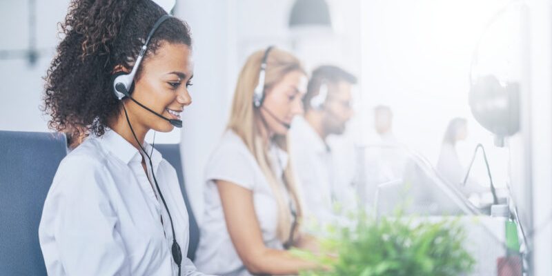 Call center worker accompanied by her team. Smiling customer support operator at work. Young employee working with a headset.